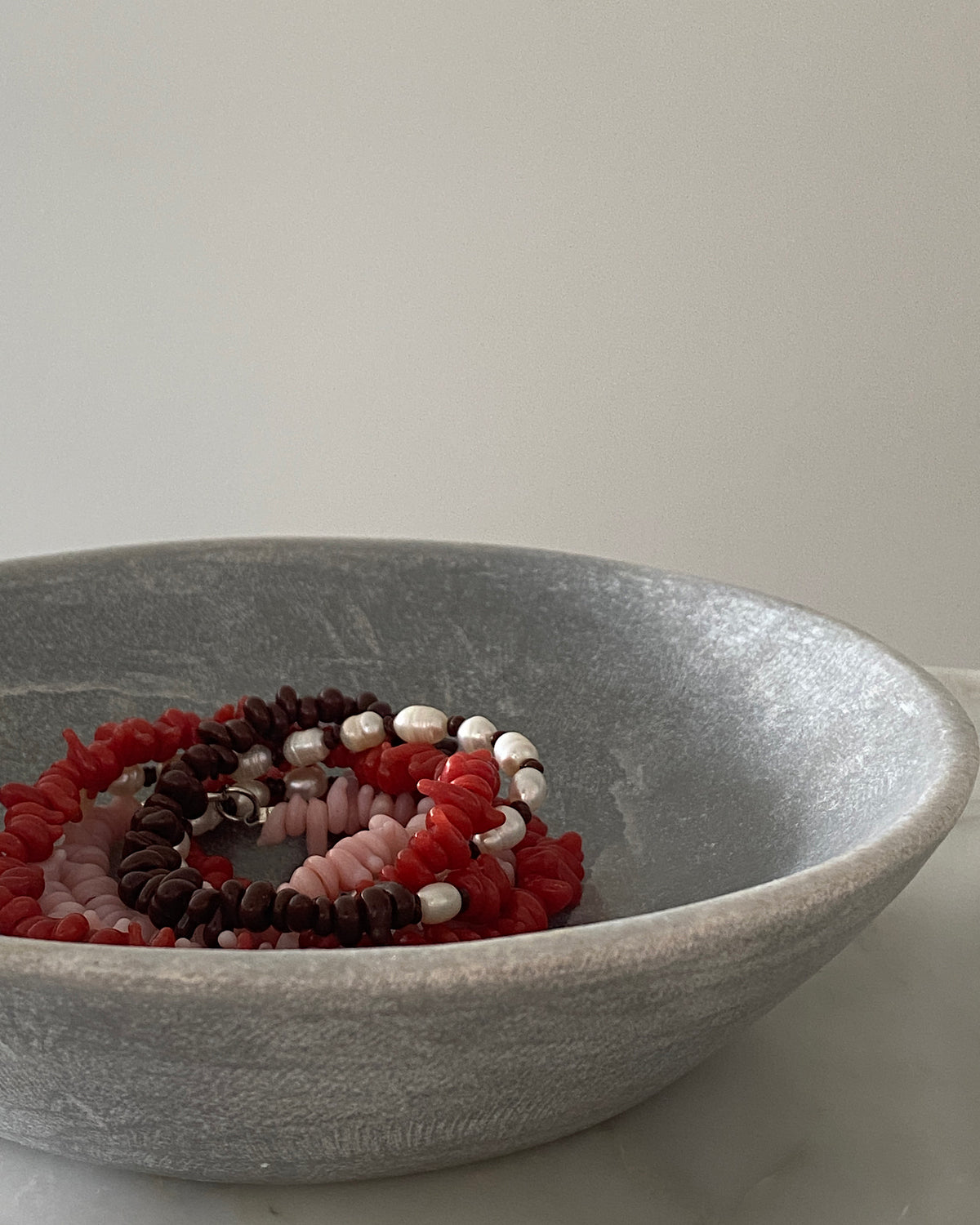 Red, pink, chocolate glass bead necklace with cream pearls sit in a bowl on marble