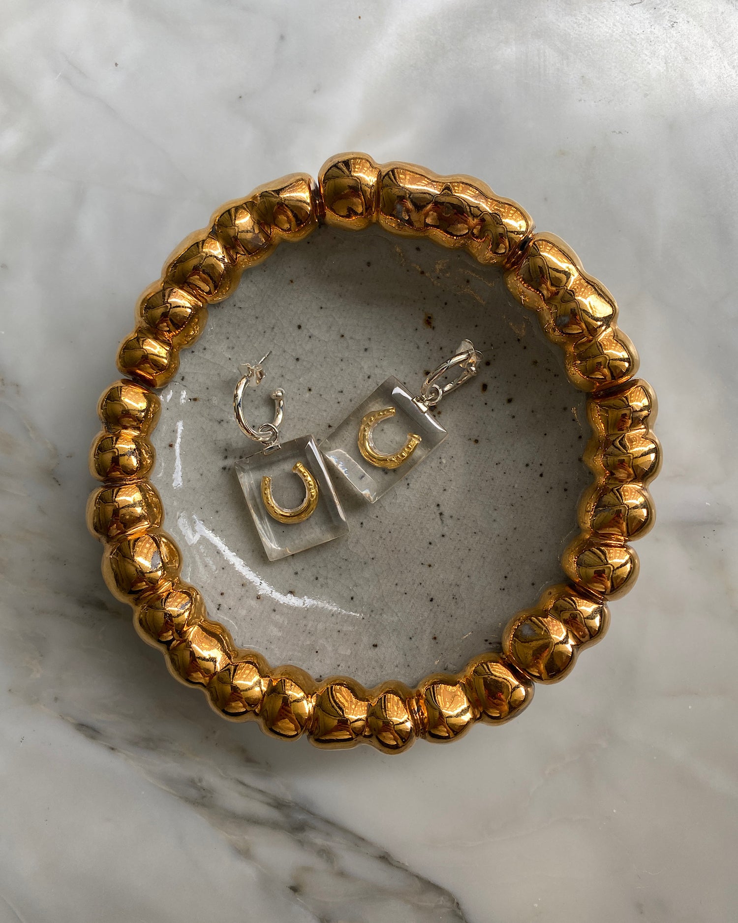 gold horse shoe and resin charm earrings with a silver hoop sit in a decorative gold bubble bowl