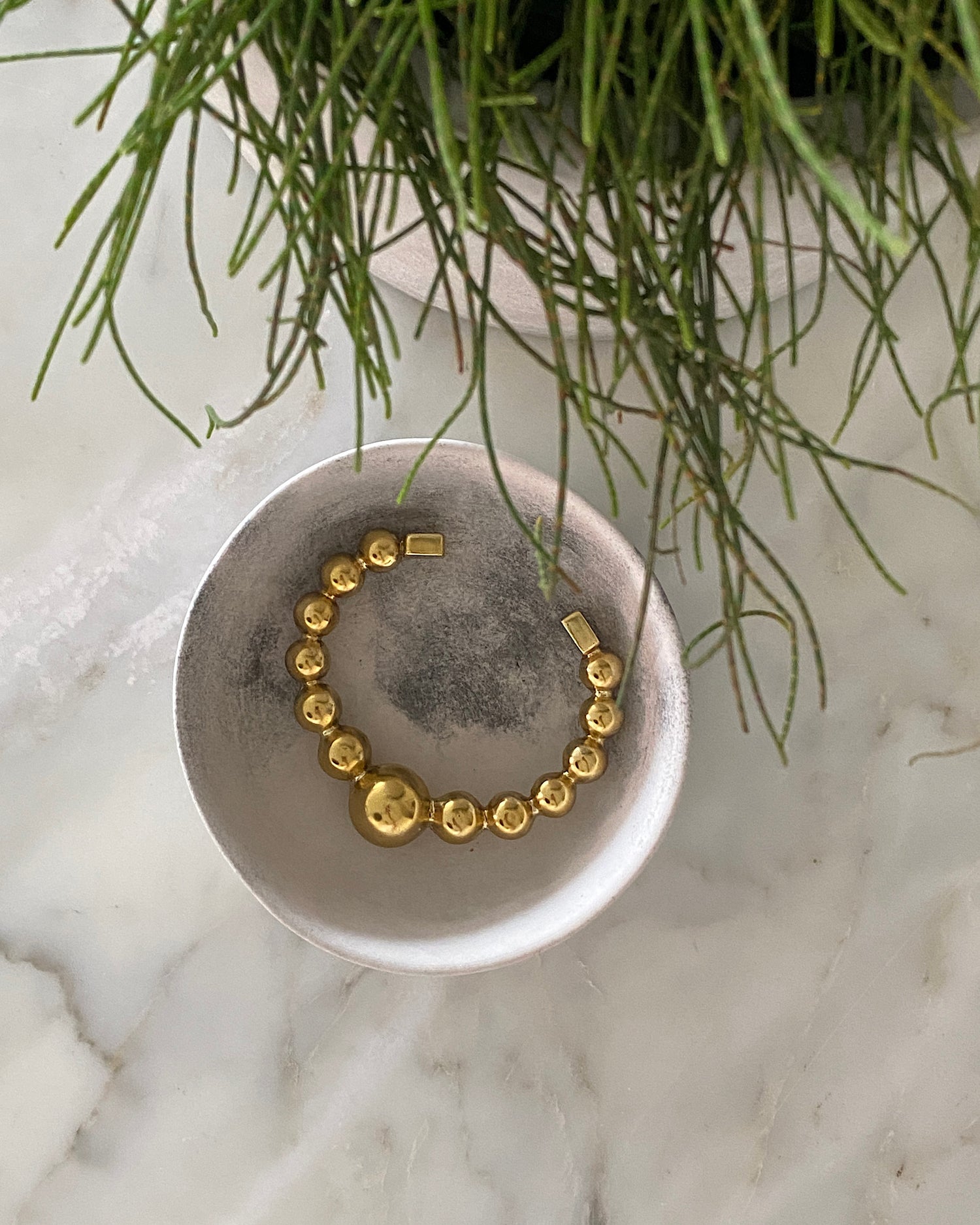 an open gold cuff bangle consisting of numerous interlinked gold balls sits in a bowl on a marble top
