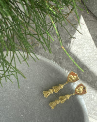 Gold ornate tassel and emu shield earrings sit in a bowl on concrete background