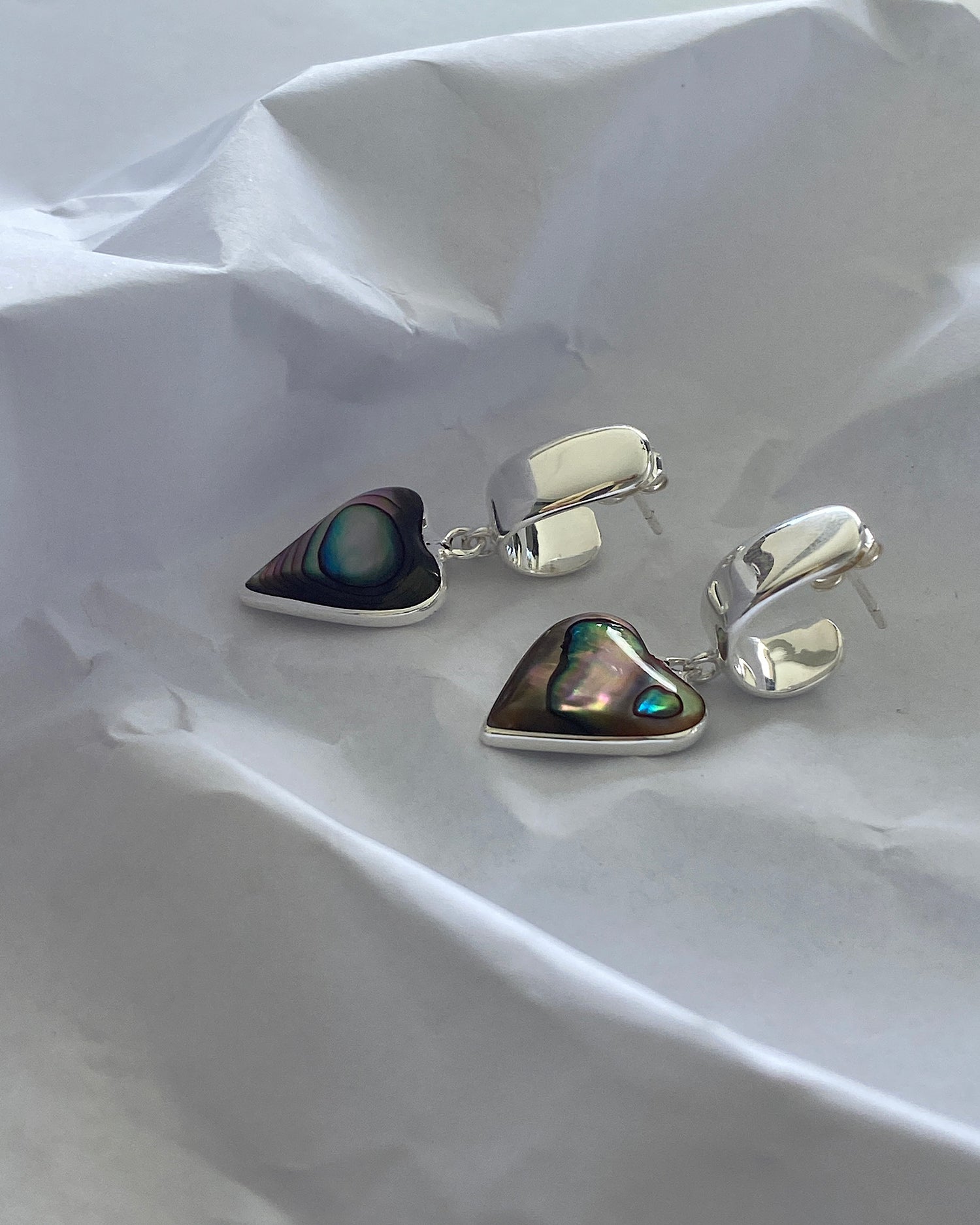 Abalone heart charms sit of a half hoop style earring on a white paper background