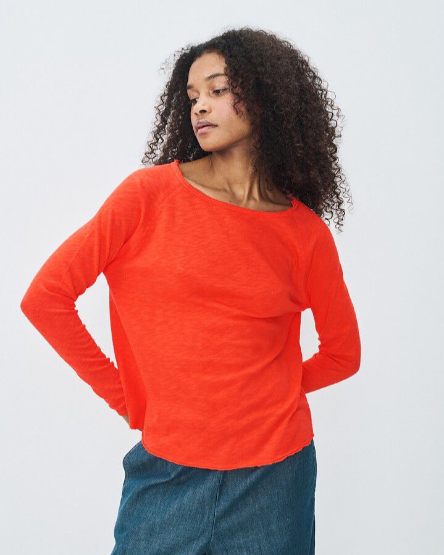 Woman wearing an orange long-sleeve boat neck top and blue jeans on a light gray background avail at The Pared Store