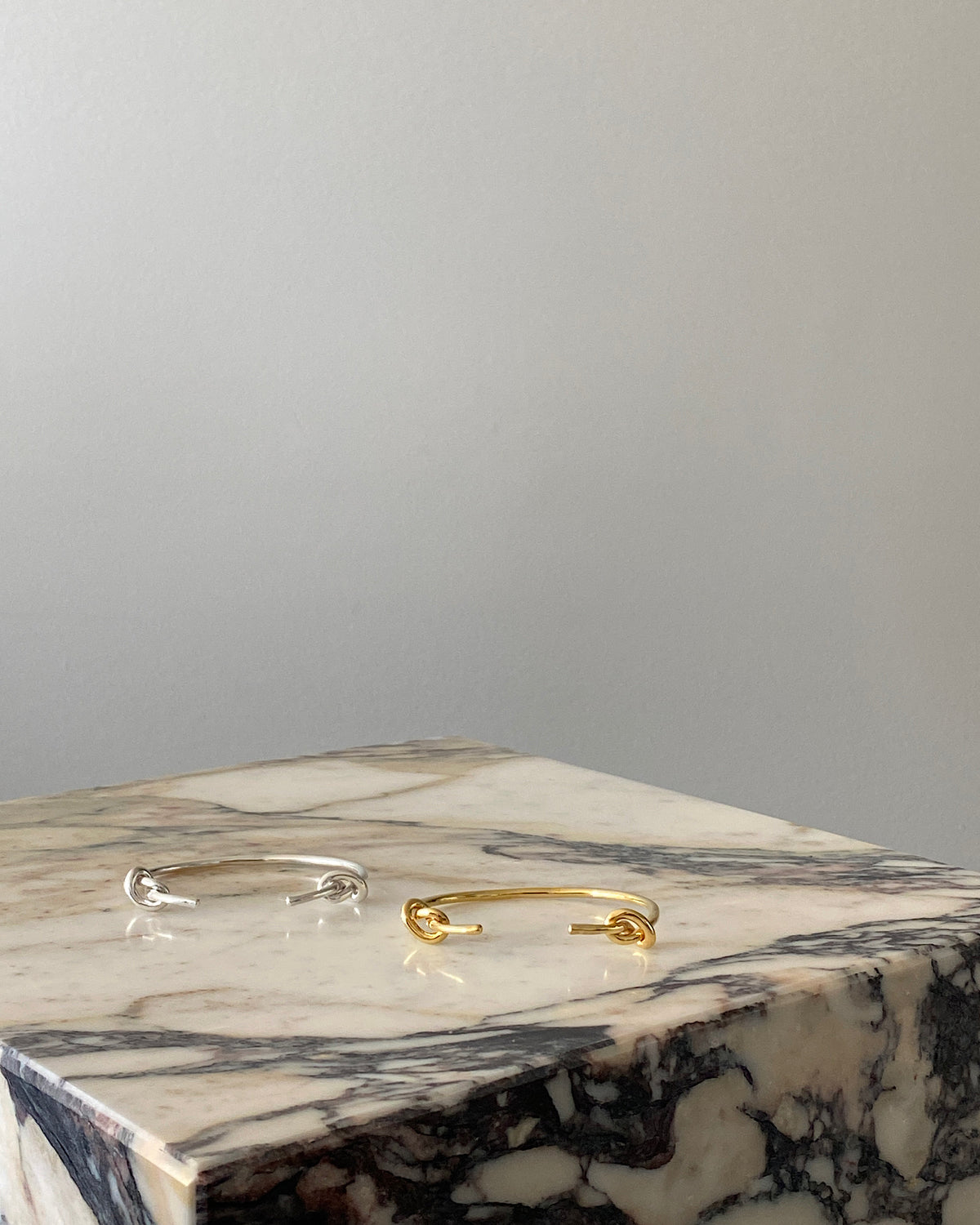 Gold and silver open cuff bracelets with knots at the end sit on a marble plinth