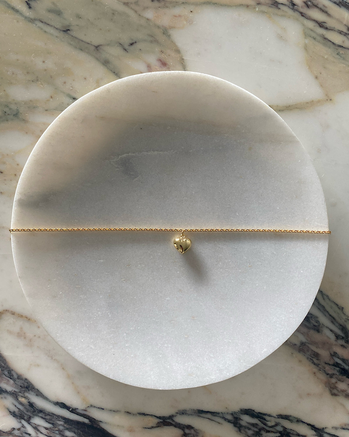 a gold heart charm on a gold chain sits on a marble plate