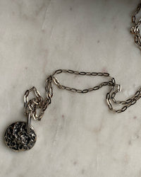 a silver necklace with a silver textured disc sits on a white marble tray