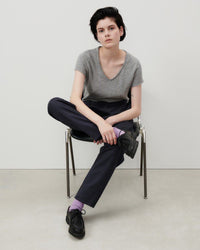 Model sits in a chair wearing a grey v neck raw edge T-shirt, black pants and shoes and lilac socks