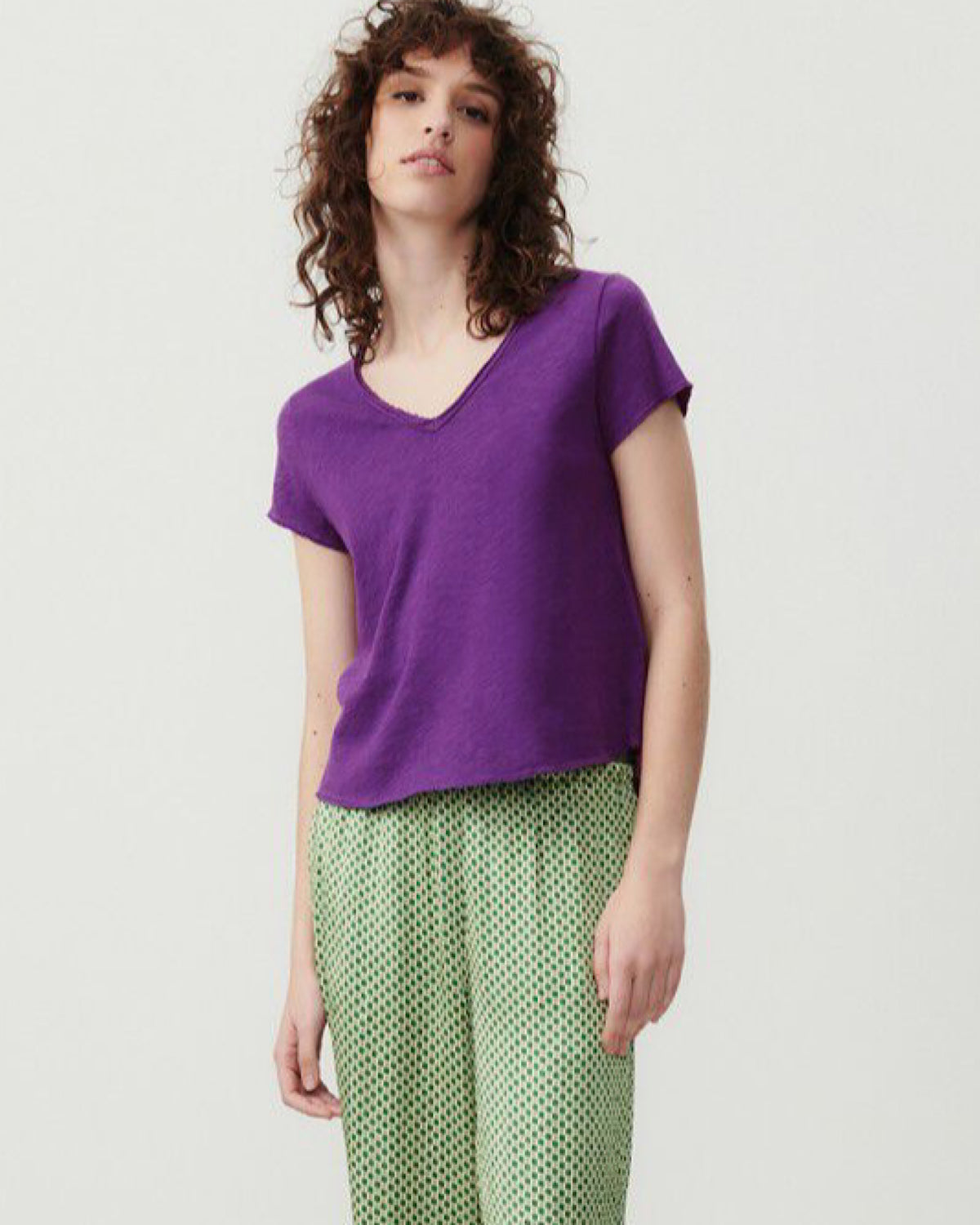 Model wears a vibrant purple v neck t-shirt from American Vintage with a raw edge finish and green patterned pants