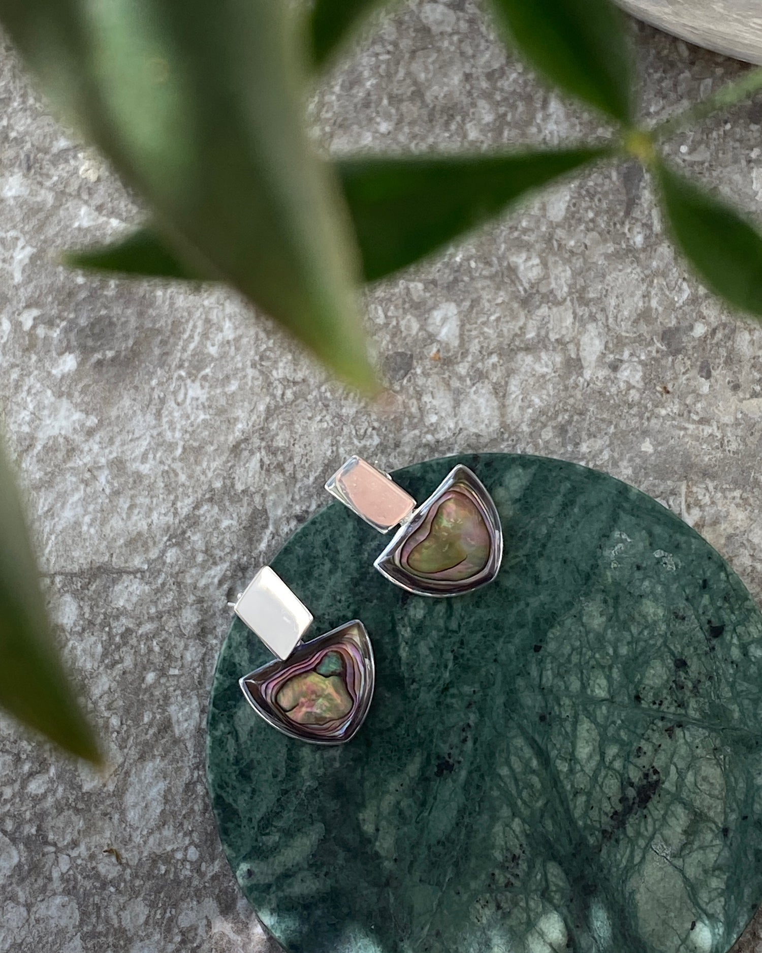 Silver and abalone earrings sit of a green marble circle on a concrete background