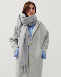 a model wears a pale grey jacket and scarf with fringing