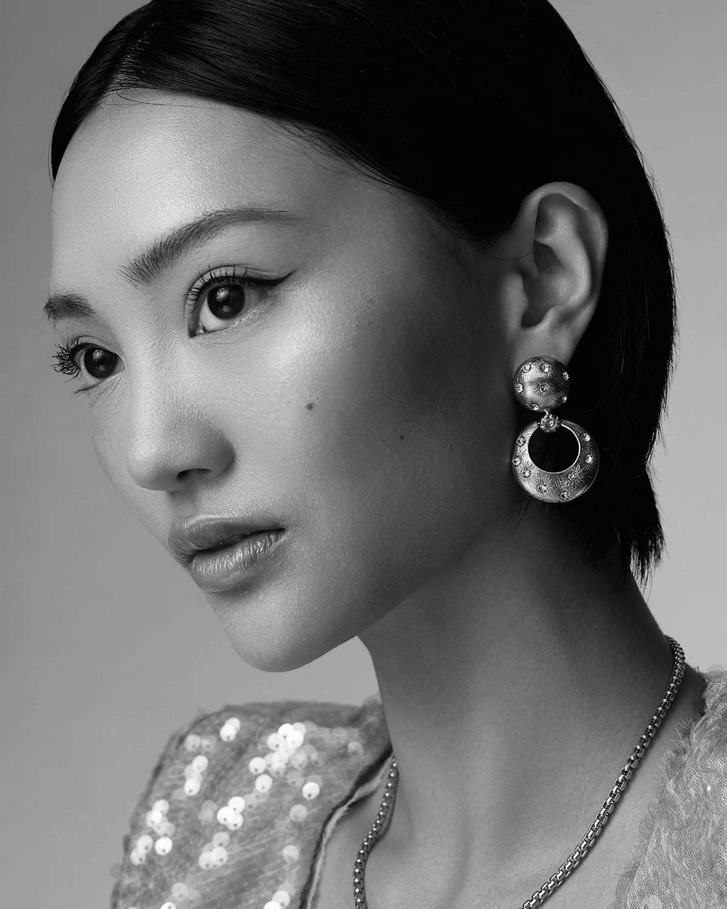 a model wears a sequin top andGold earrings featuring a round stud top and open circle drop inlaid with crystals