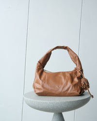 a tan coloured slouchy bag with handle and tassel sits on a stool