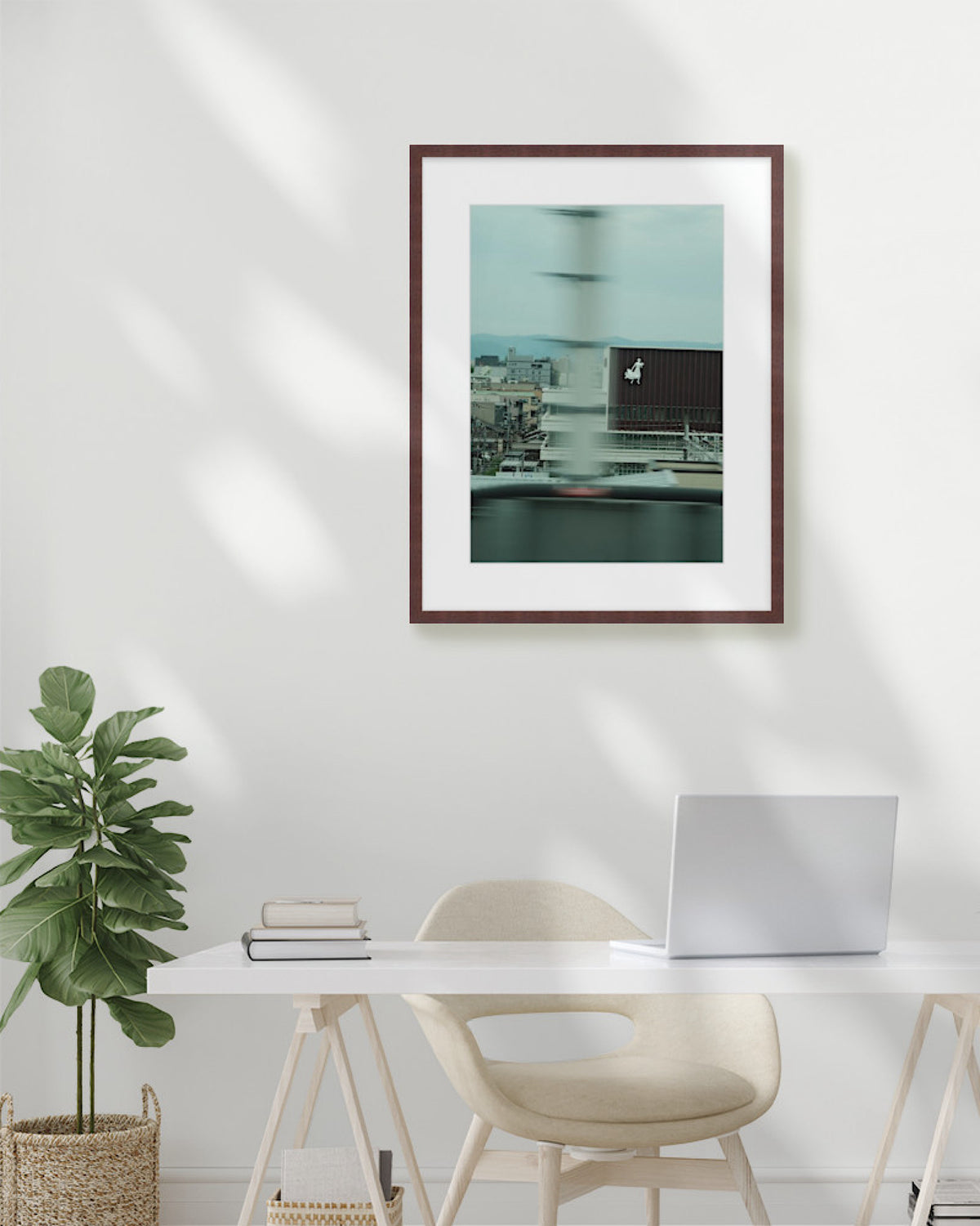 Framed artwork above a desk with a chair and plant in a minimalistic room.