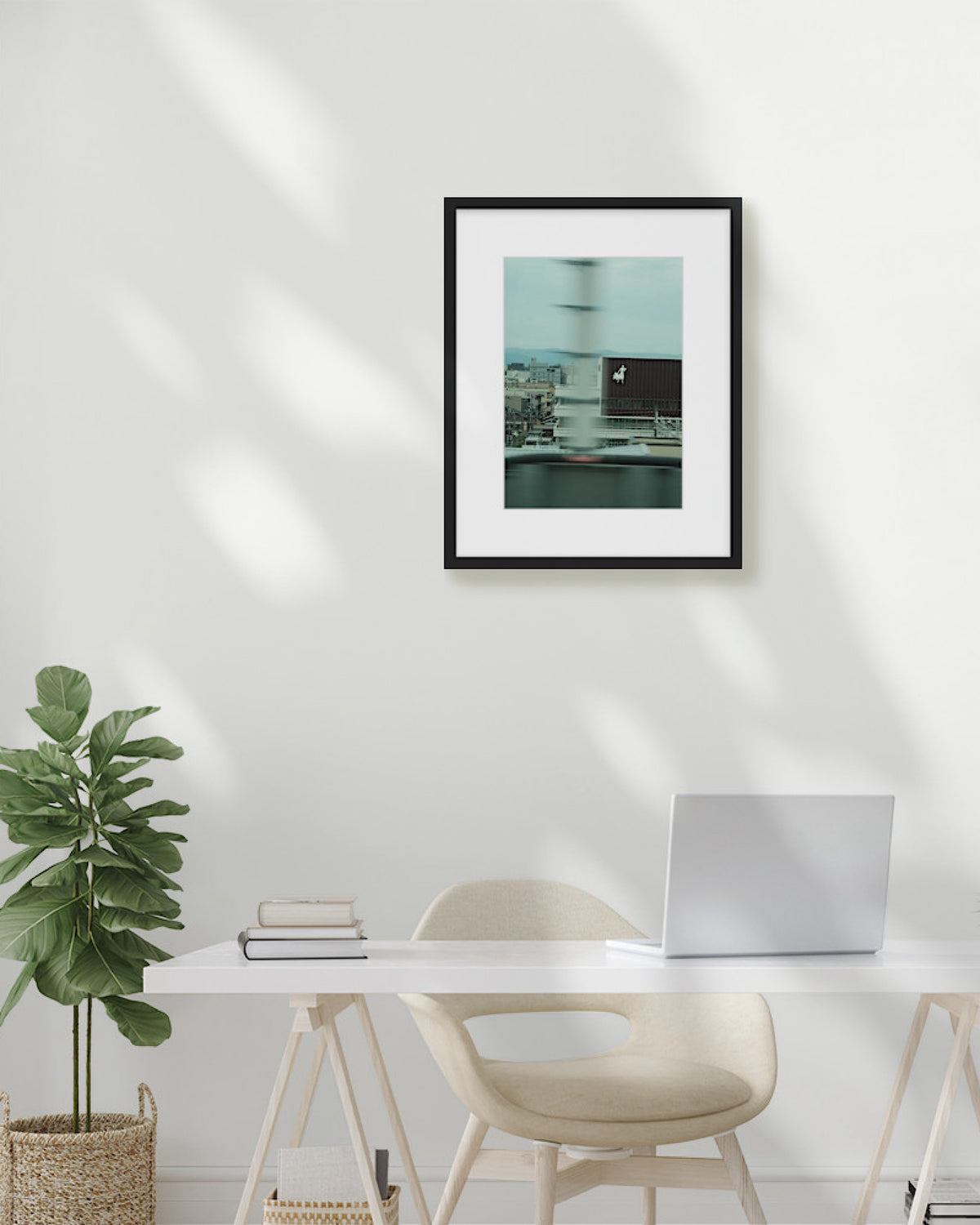 Framed artwork on a wall above a desk with a plant and laptop.