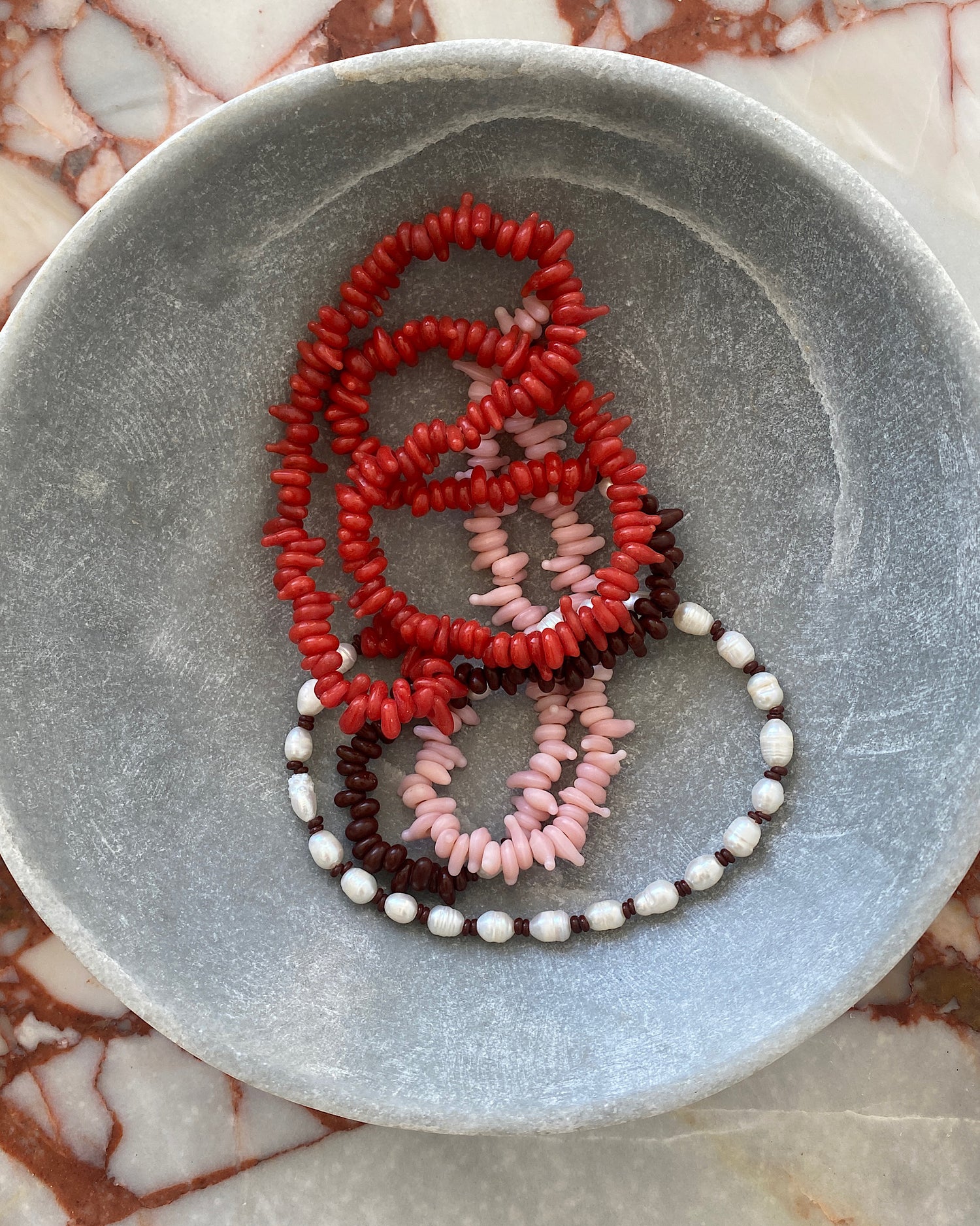 Red, pink, chocolate glass bead necklace with cream pearls sit in a bowl on marble