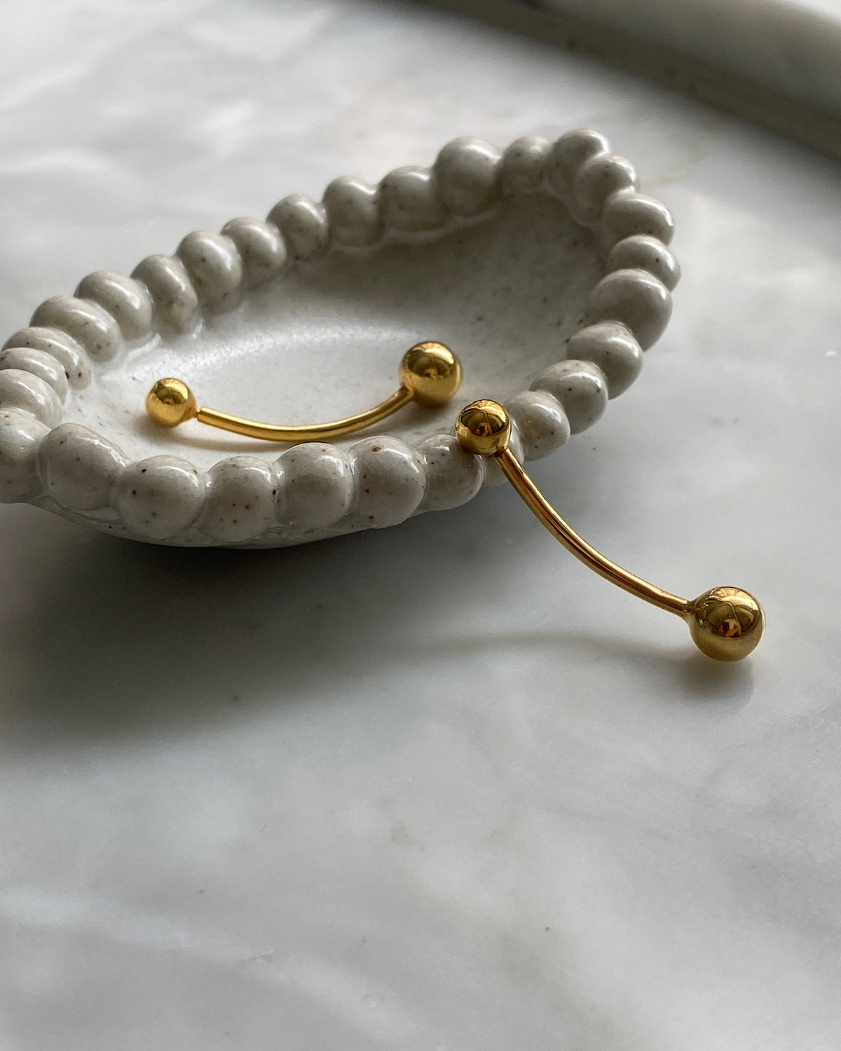 Gold double ended ball earrings sit in a cream dish with a border of self balls