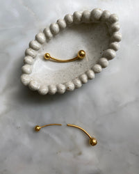 Gold double ended ball earrings sit in a cream dish with a border of self balls