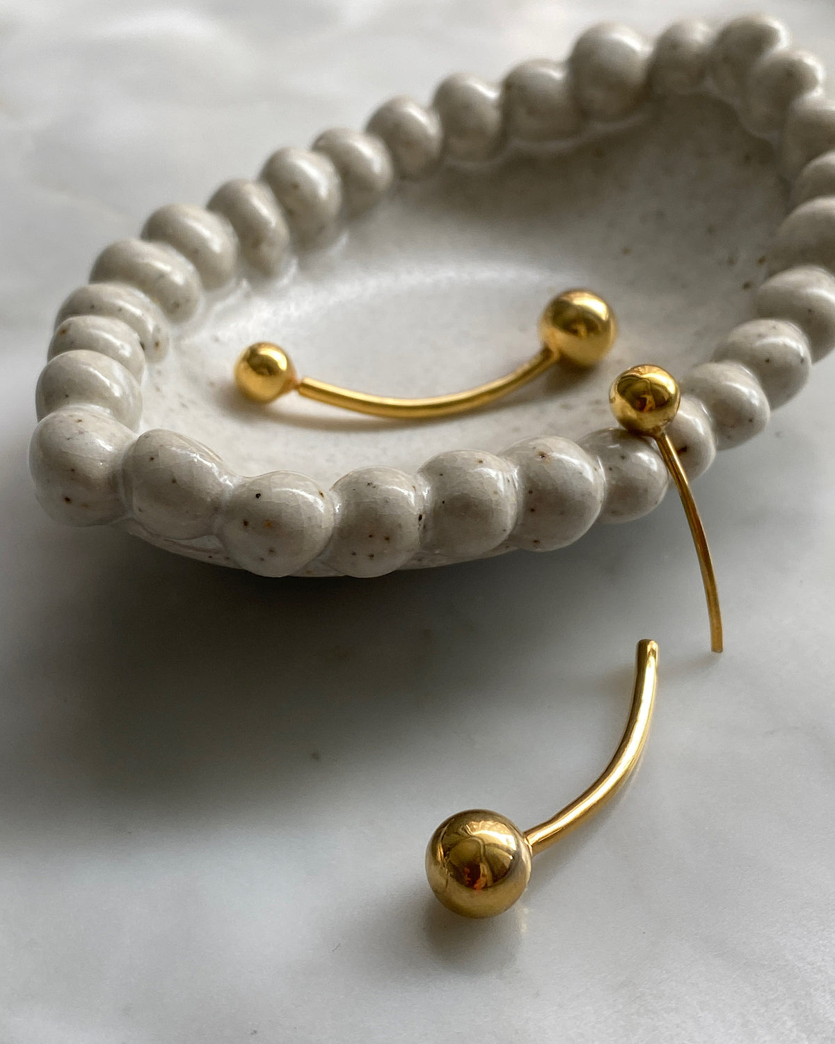 Gold double ended ball earrings sit in a cream dish with a border of self balls