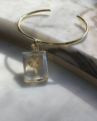 A clear resin rectangular charm with a gold four leaf clover and silver clasp is attached to a gold bangle