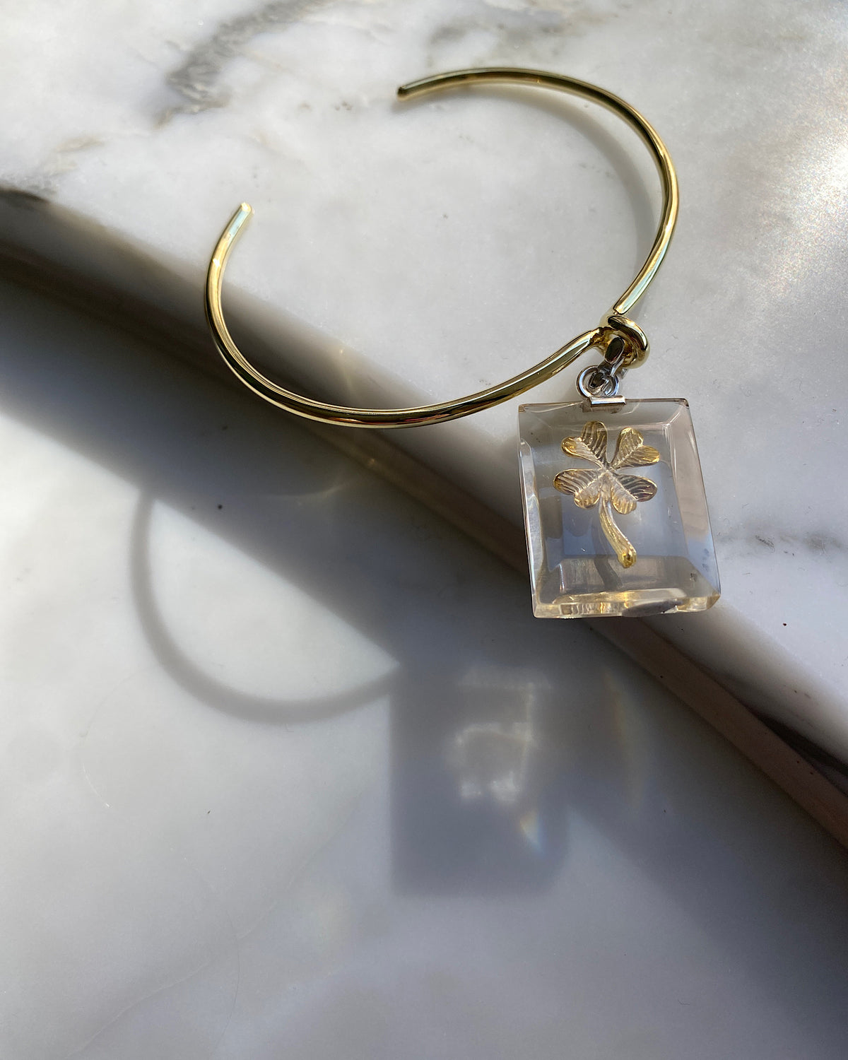 A clear resin rectangular charm with a gold four leaf clover and silver clasp sits attached to a gold bangle