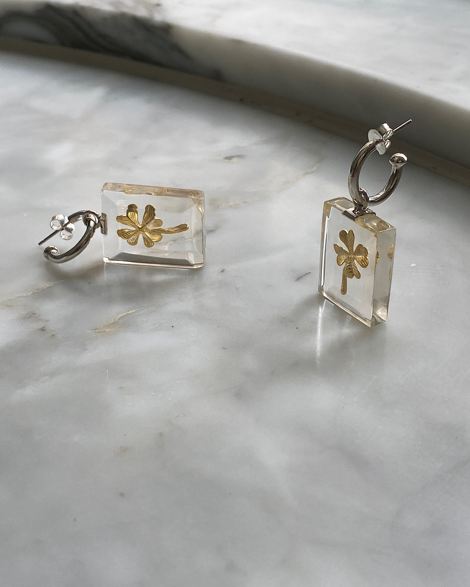 Clear resin rectangles with a gold four leaf charm hang from a silver hoop earring against a marble background