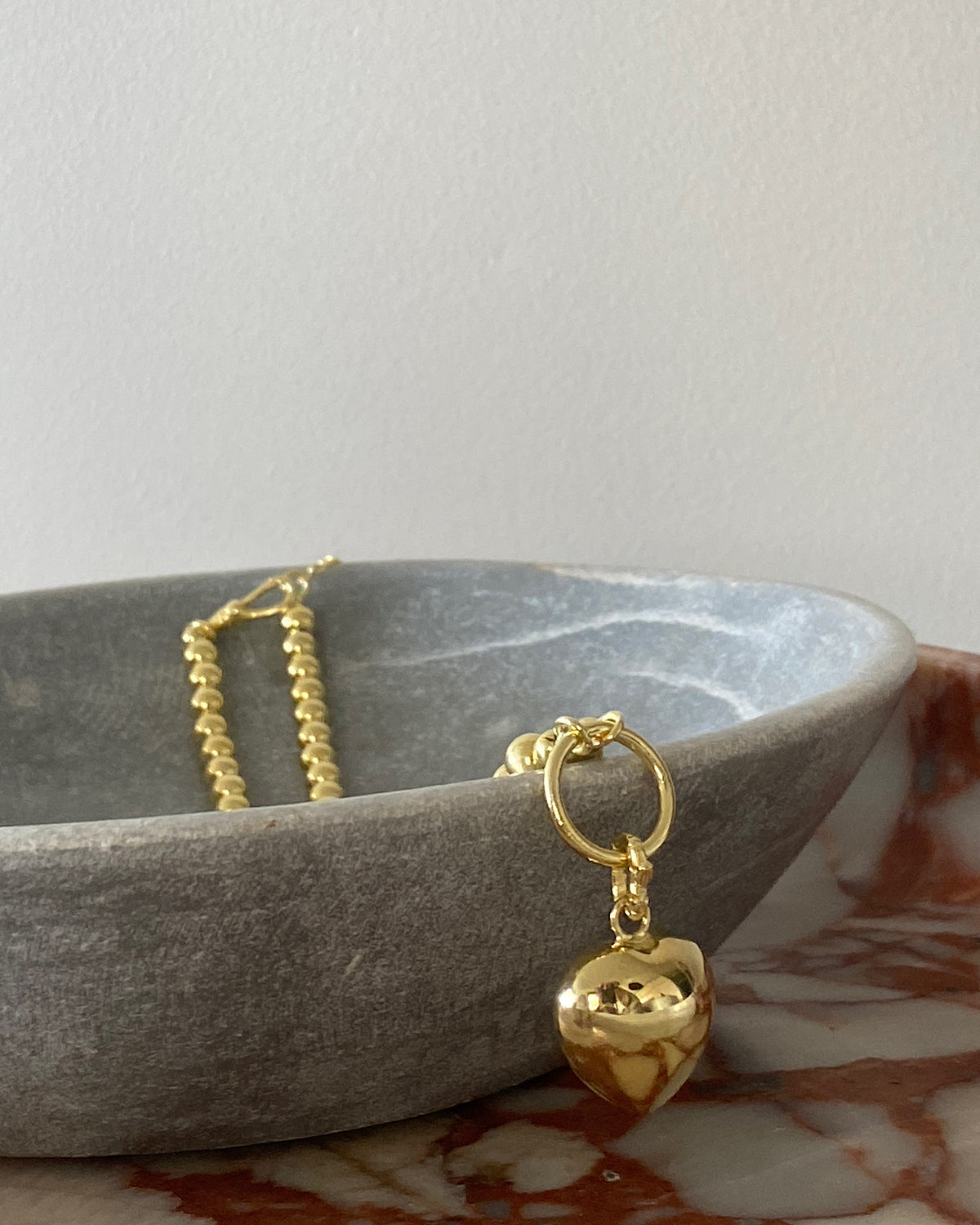 a gold ball chain with a central ring for hanging charms with a charm heart charm attached in a bowl on marble