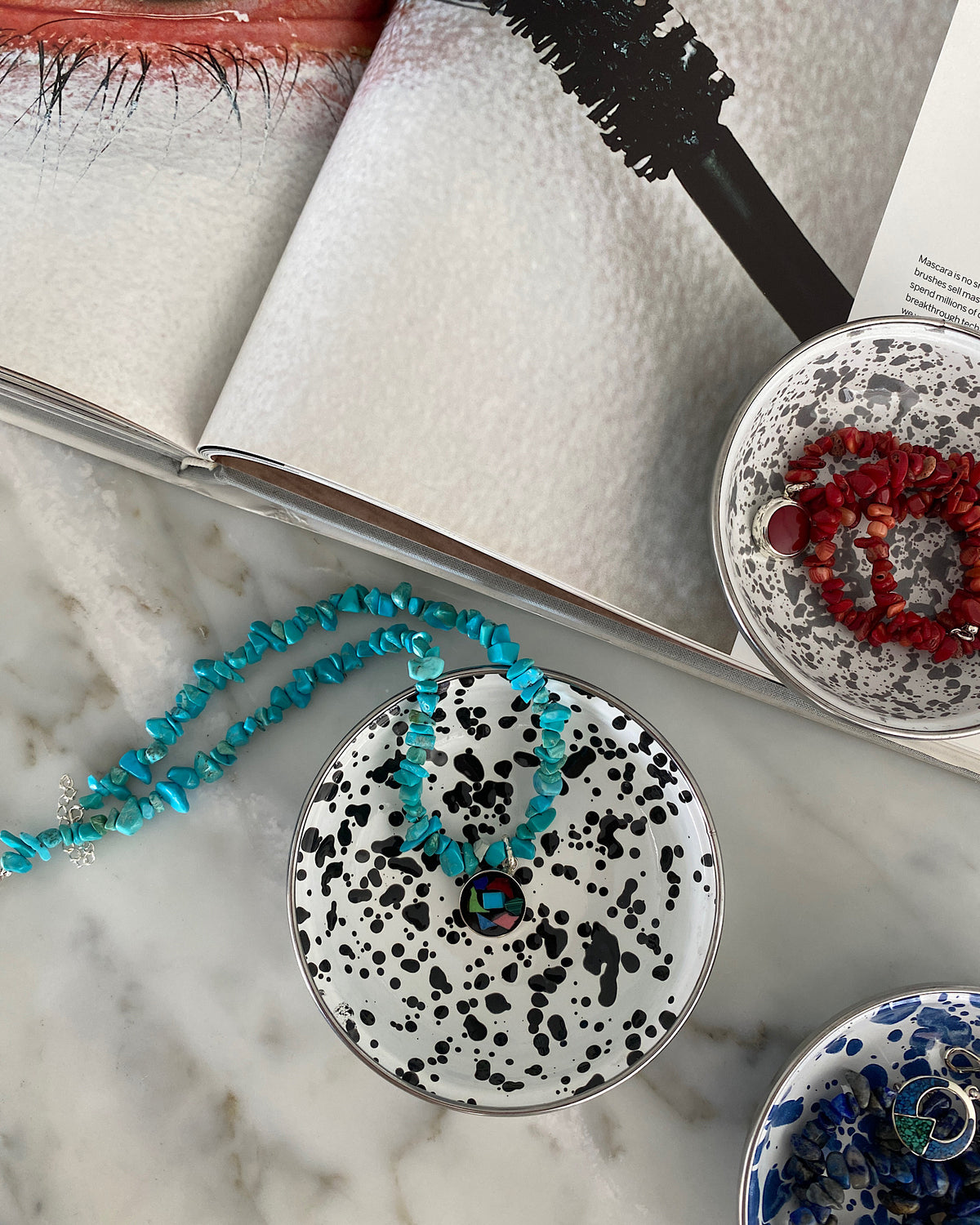 a turquoise jagged necklace with a central round multi colour and silver pendant sits in a bowl alongside other necklaces