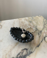 Simple silver oval chain bracelet with a clasp has a heart charm added sits in a black bowl on a marble base