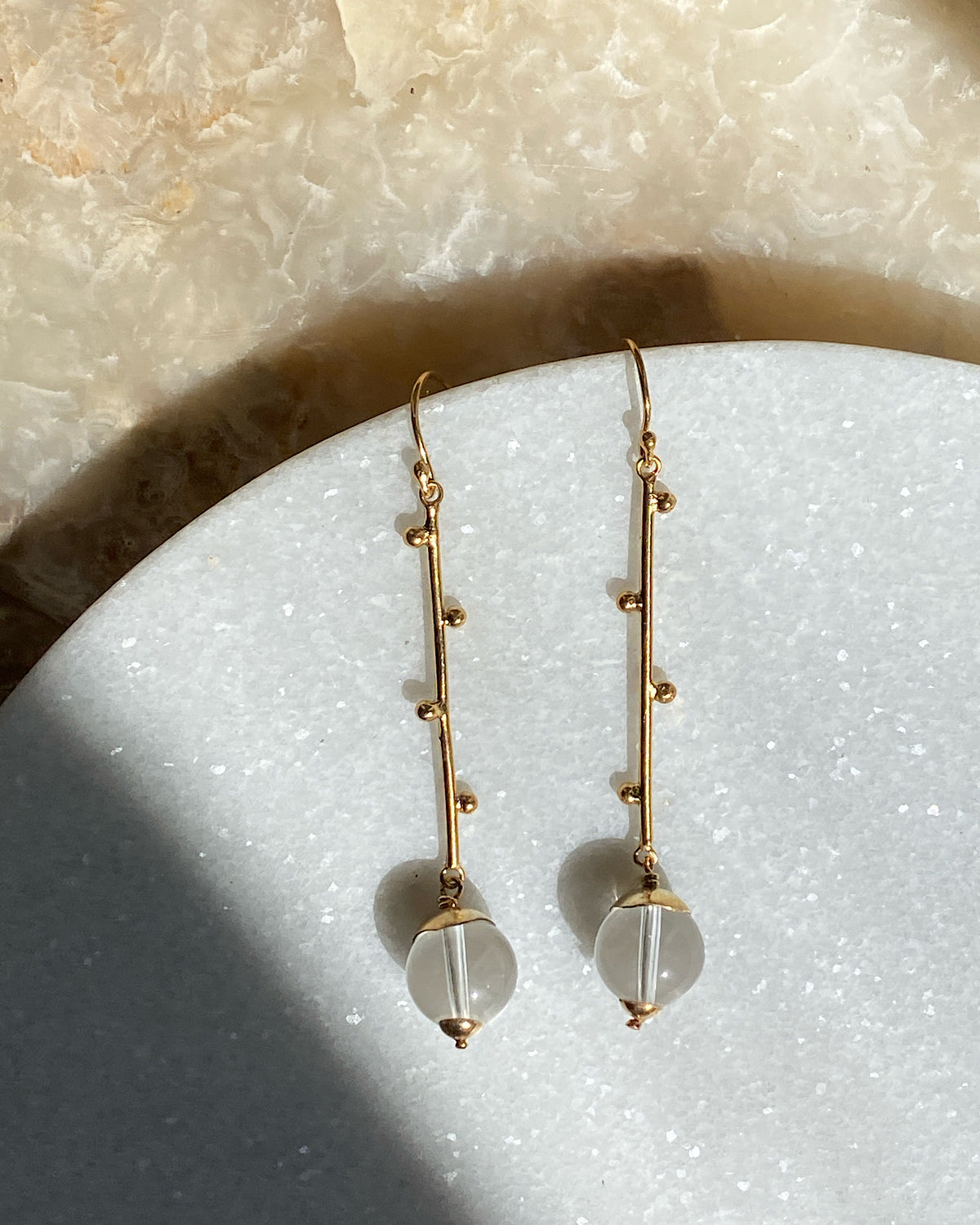 Long gold drop earrings with clear quartz sit on a marble plate