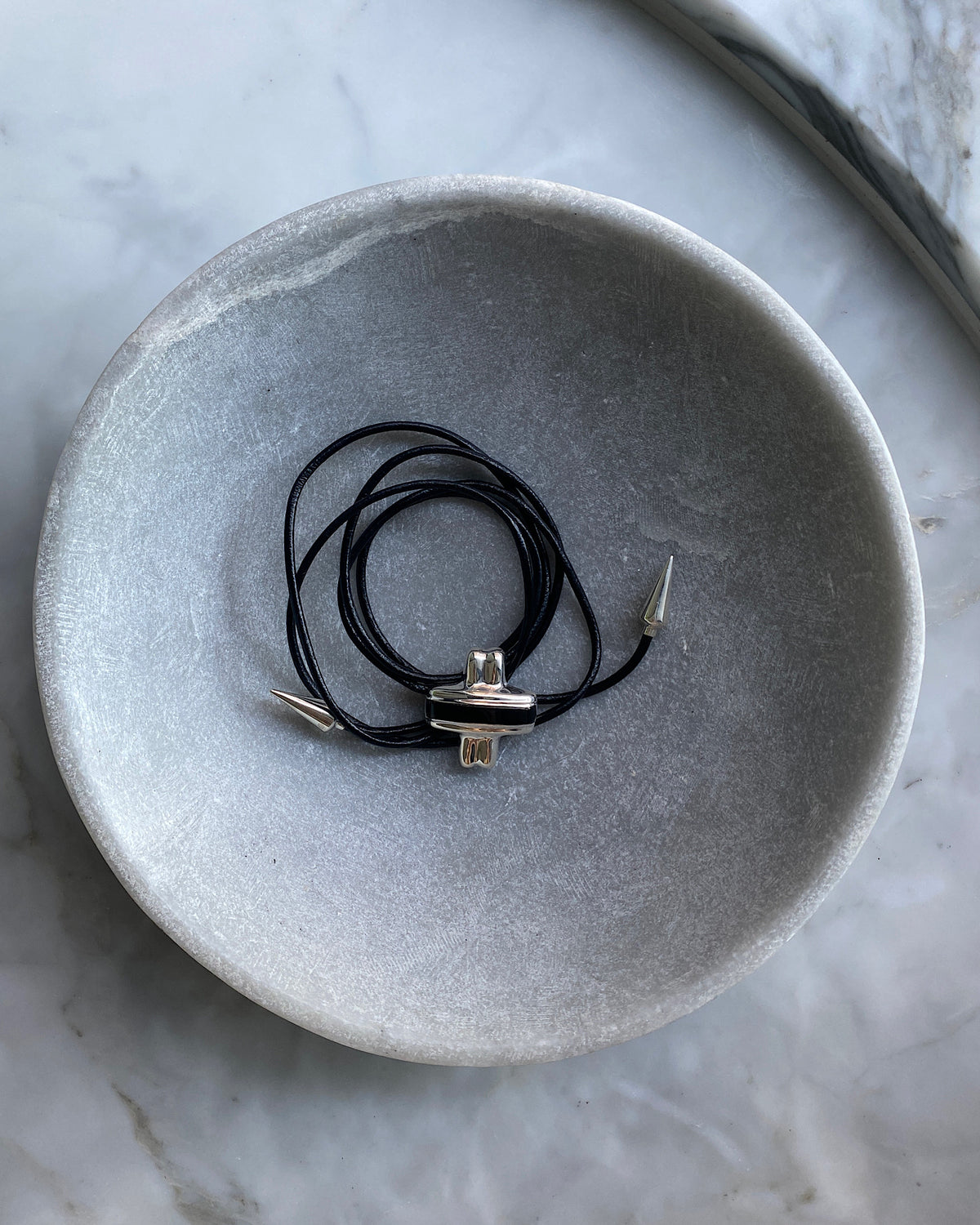 Silver and black cross pendant on a black leather tie with silver pointed ends sits in a bowl