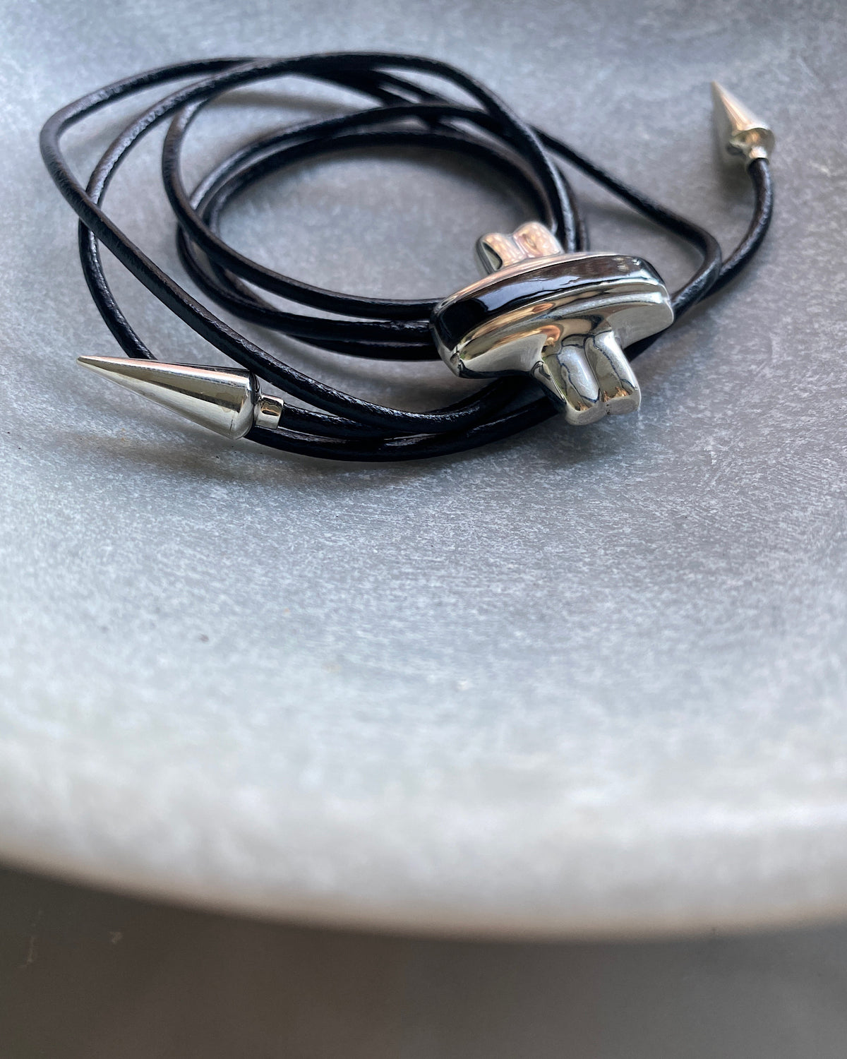 Silver and black cross pendant on a black leather tie with silver pointed ends sits in a bowl