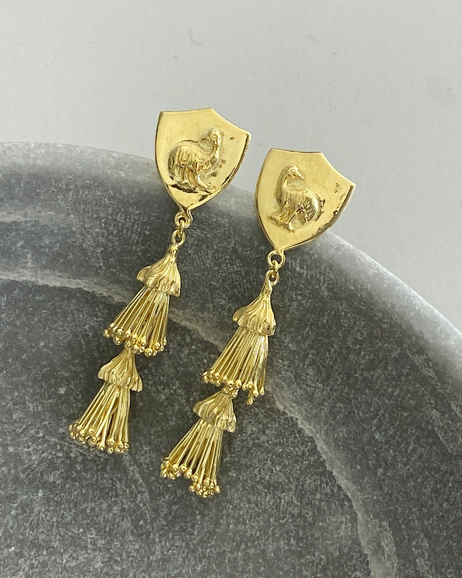 Gold ornate tassel and emu shield earrings sit in a bowl on concrete background