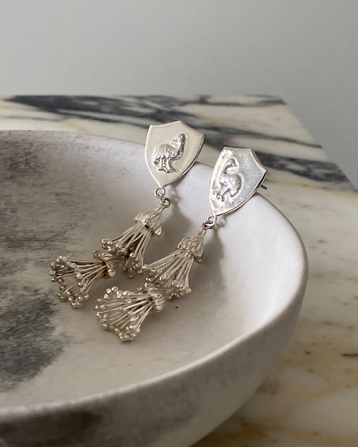 Silver ornate tassel and emu shield earrings sit in a bowl on marble background