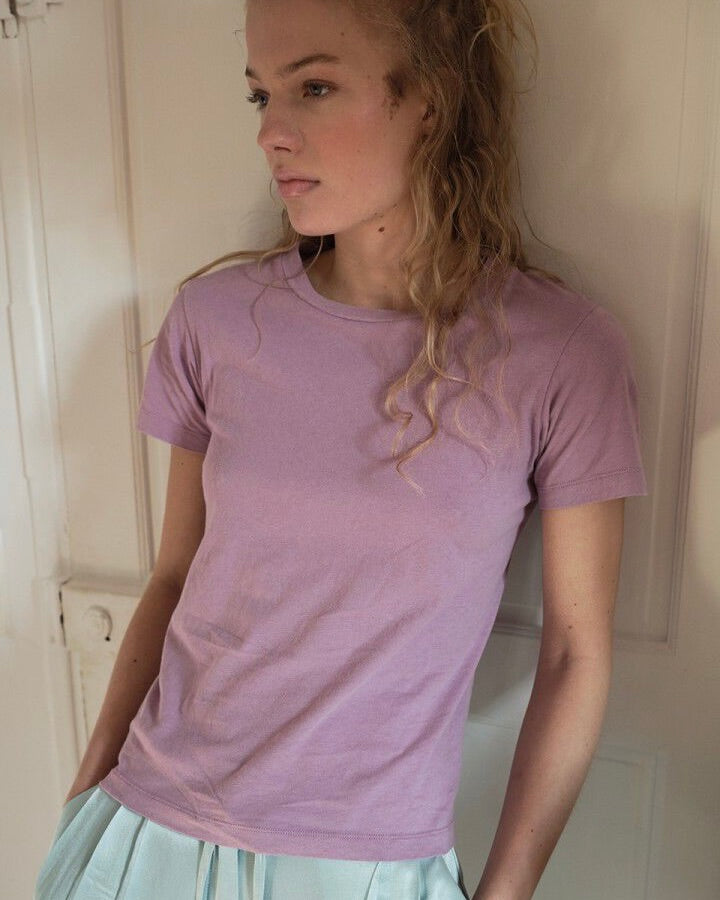 Woman wearing a purple t-shirt standing against a plain wall.