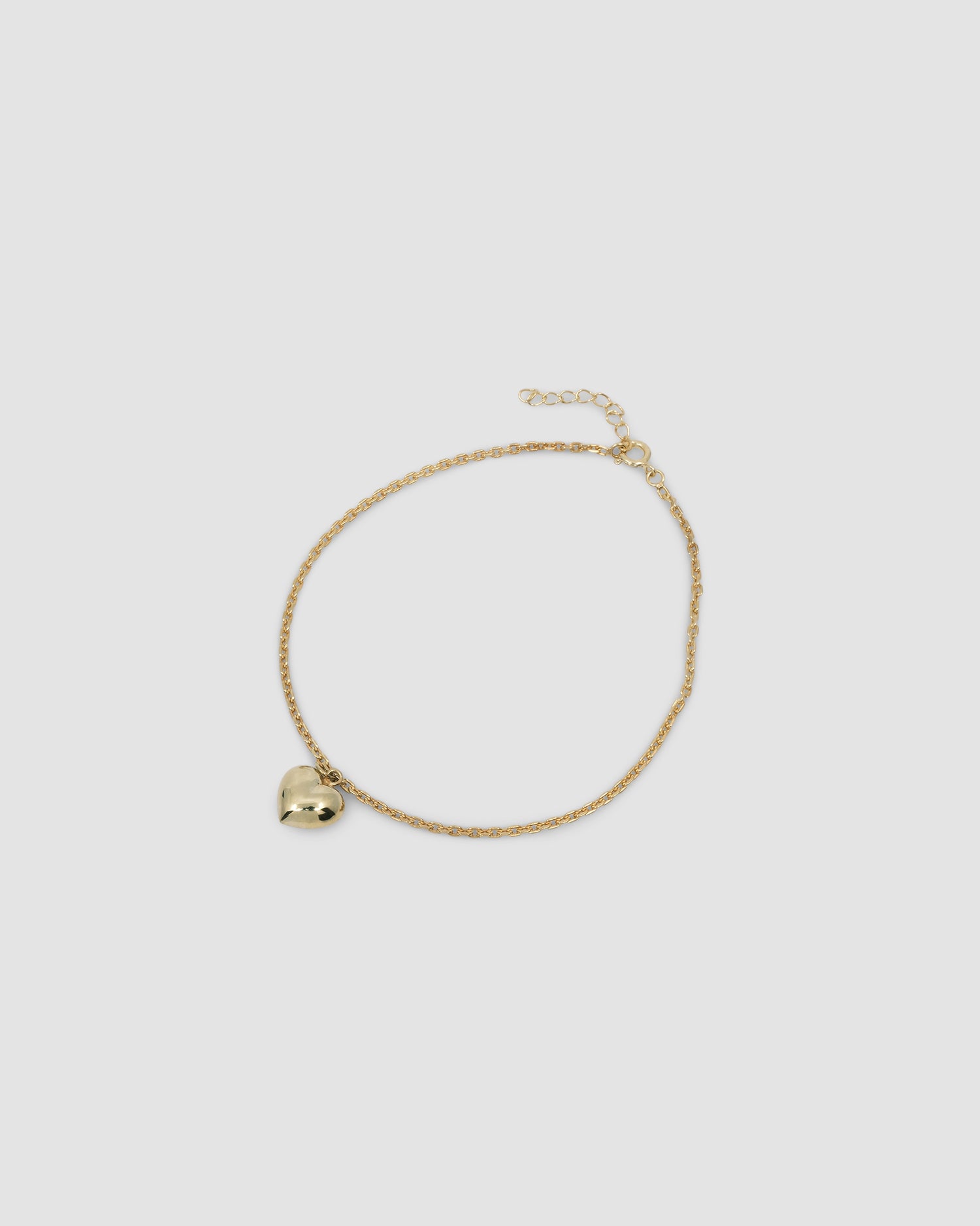 A fine gold chain anklet with a single heart charm sits on a grey background