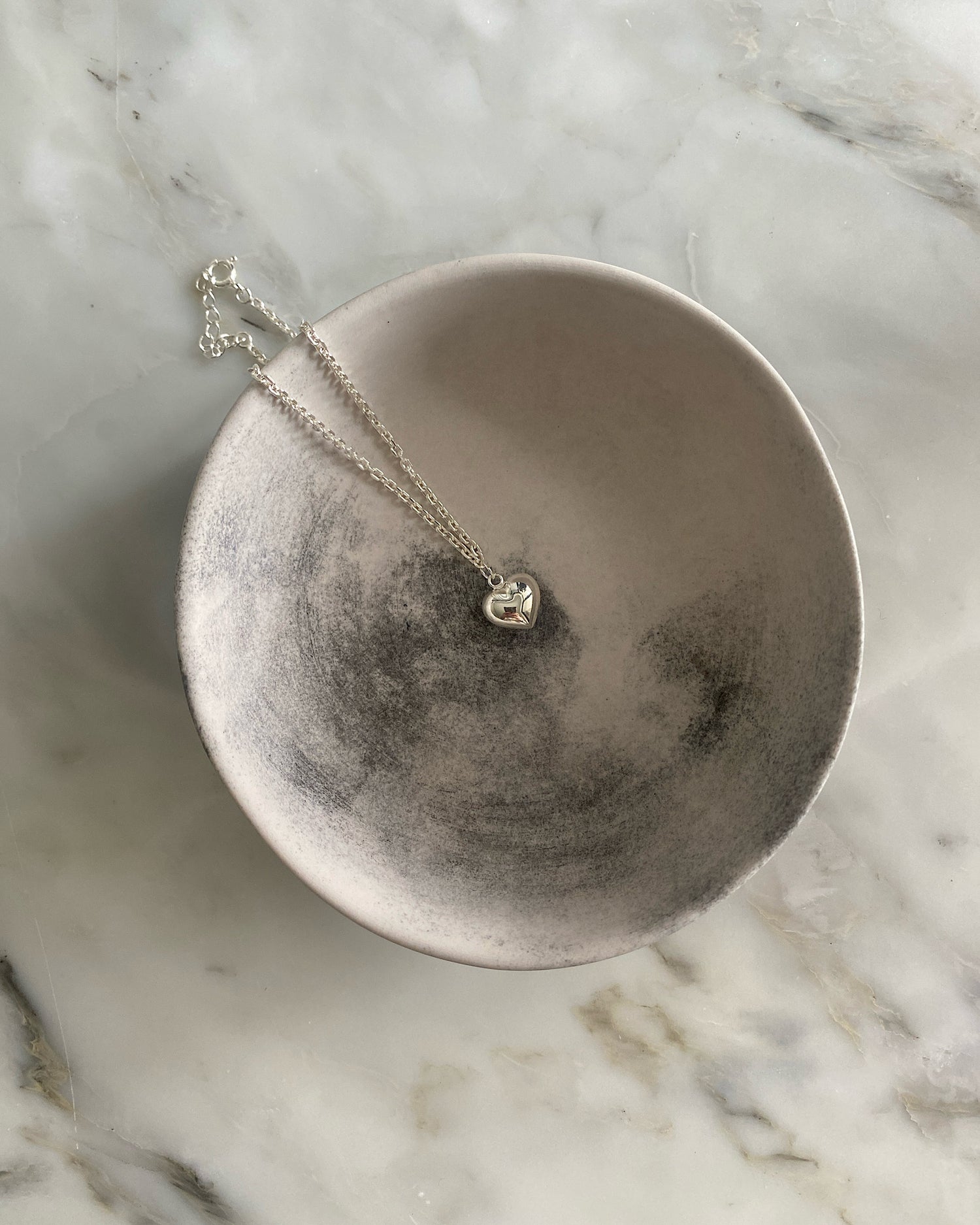 a sterling silver anklet with a heart charm sits in a bowl on a marble table