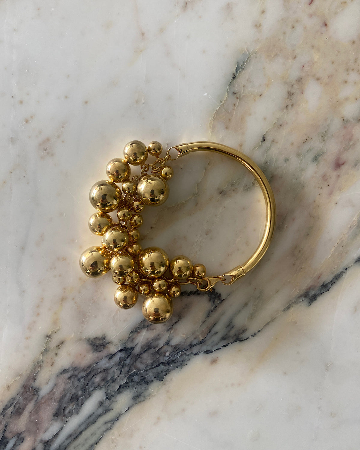 a gold bangle with a group of various shaped gold balls attached to half sits against a marble background