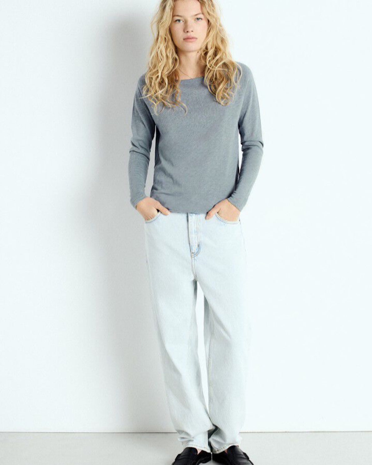 Woman wearing a grey blue boat neck top and light blue pants against a white background